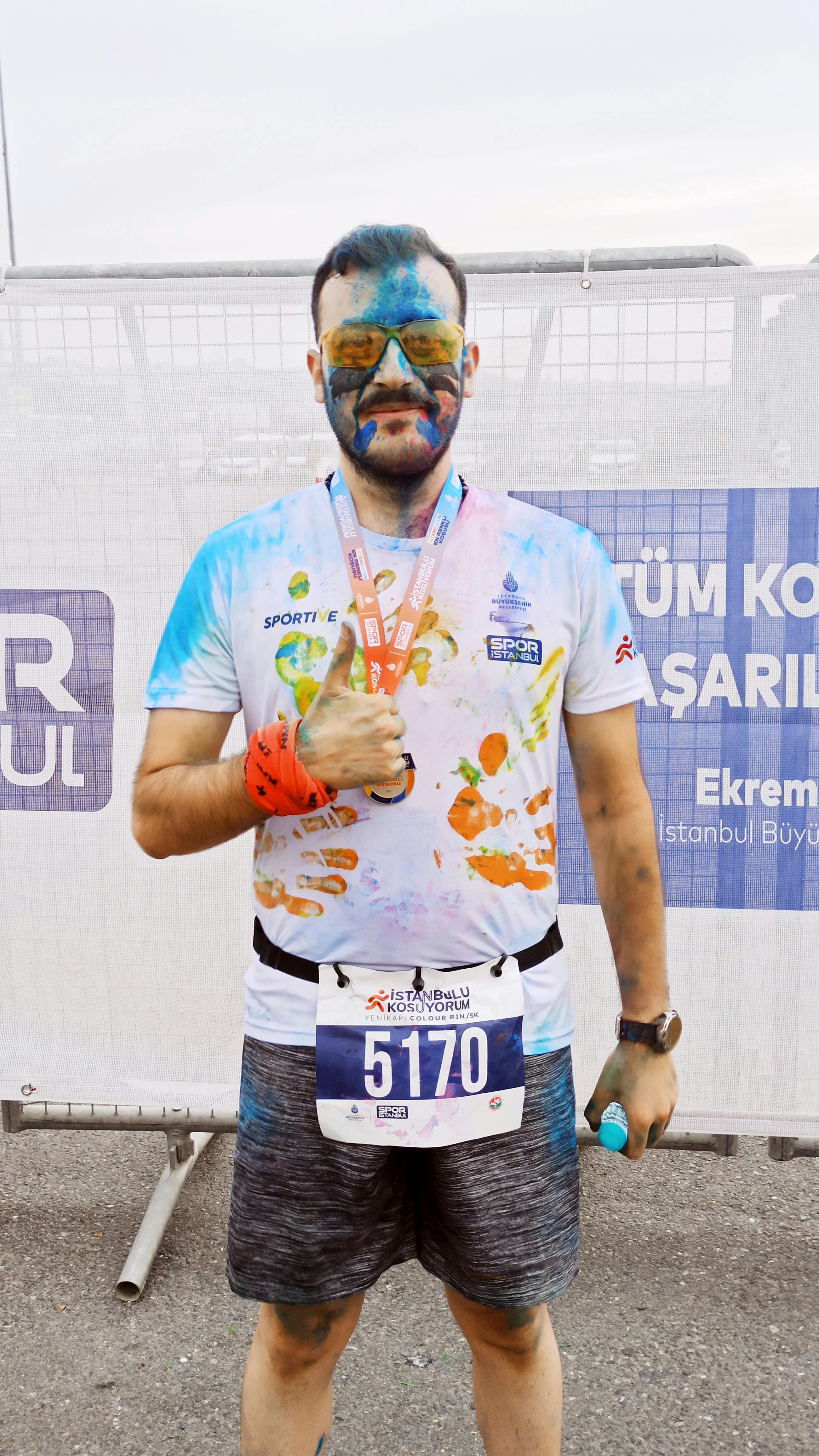 A man celebrates after a colorful fun run event, proudly displaying his medal and vibrant face paint.