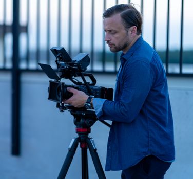 A male filmmaker in Chicago adjusting a video camera on a tripod, outdoors at twilight.