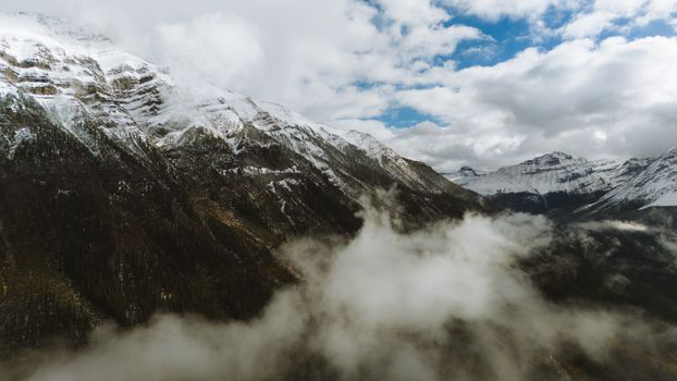 A breathtaking aerial view of snow-capped Rocky Mountains surrounded by misty clouds and blue sky.