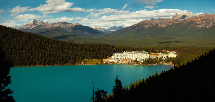 Panoramic view of a lake, forest, and hotel nestled among mountains under a clear sky.