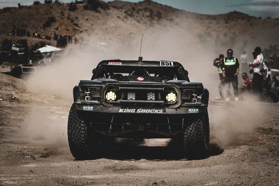 High-speed off-road racing action in the Mexicali desert with dust clouds and spectators.