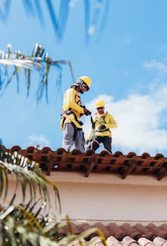 Two construction workers in safety gear working on a tiled roof under a clear blue sky.