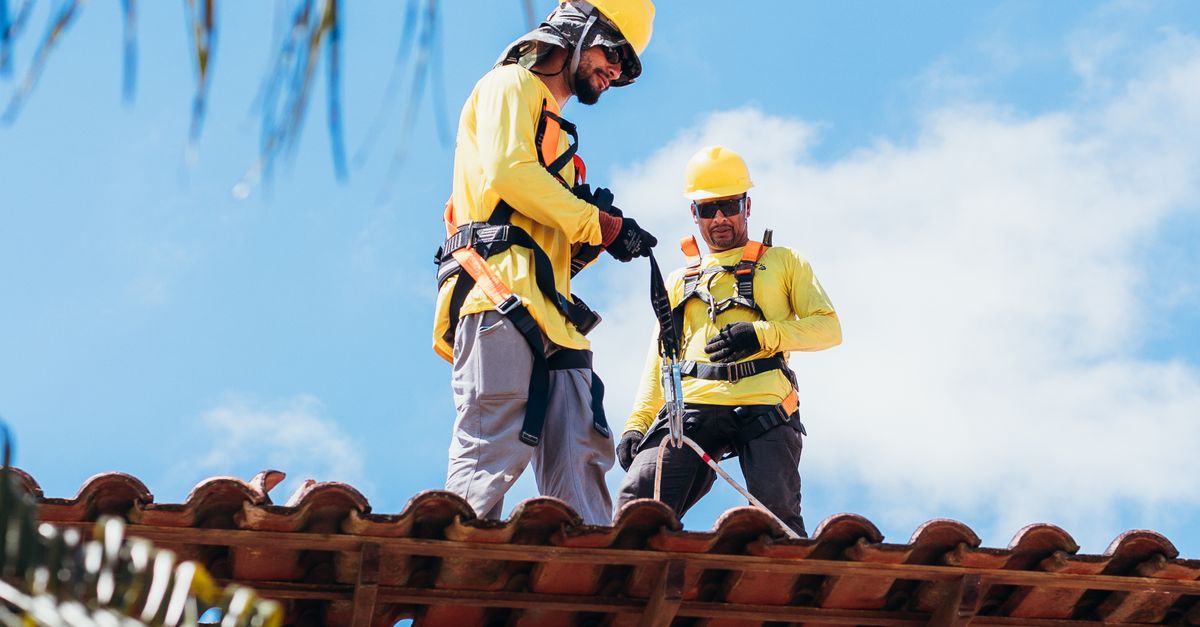 Two construction workers in safety gear working on a tiled roof under a clear blue sky.