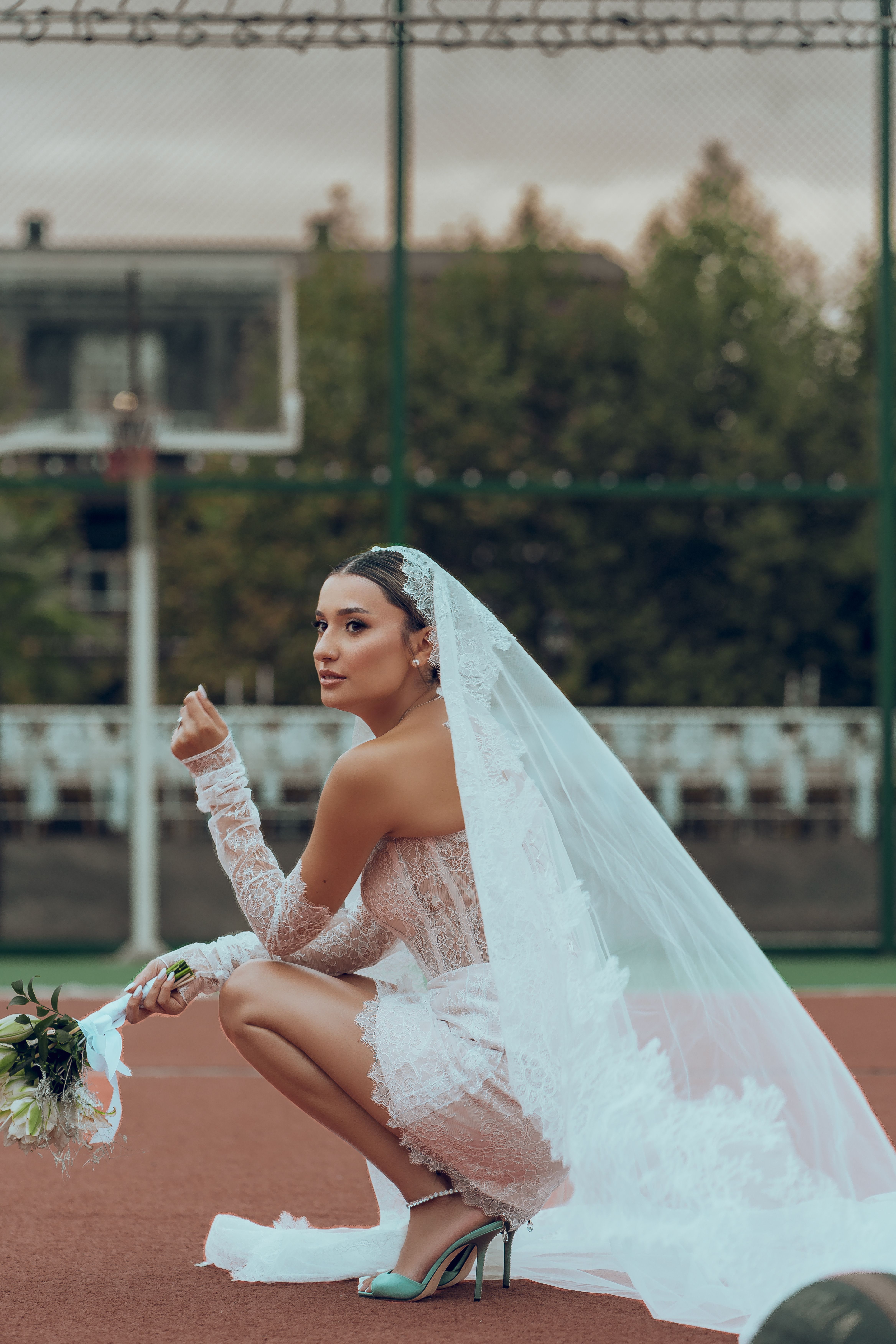 Bride in elegant dress and veil posing on tennis court with bouquet.