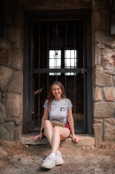 Smiling young woman sits casually by a rustic stone doorway enjoying outdoors.