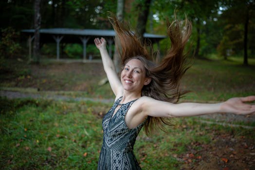 A happy woman dancing outdoors with her hair flying in a grassy forest setting.