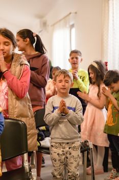 Children gathered in prayer with hands clasped at an indoor event.