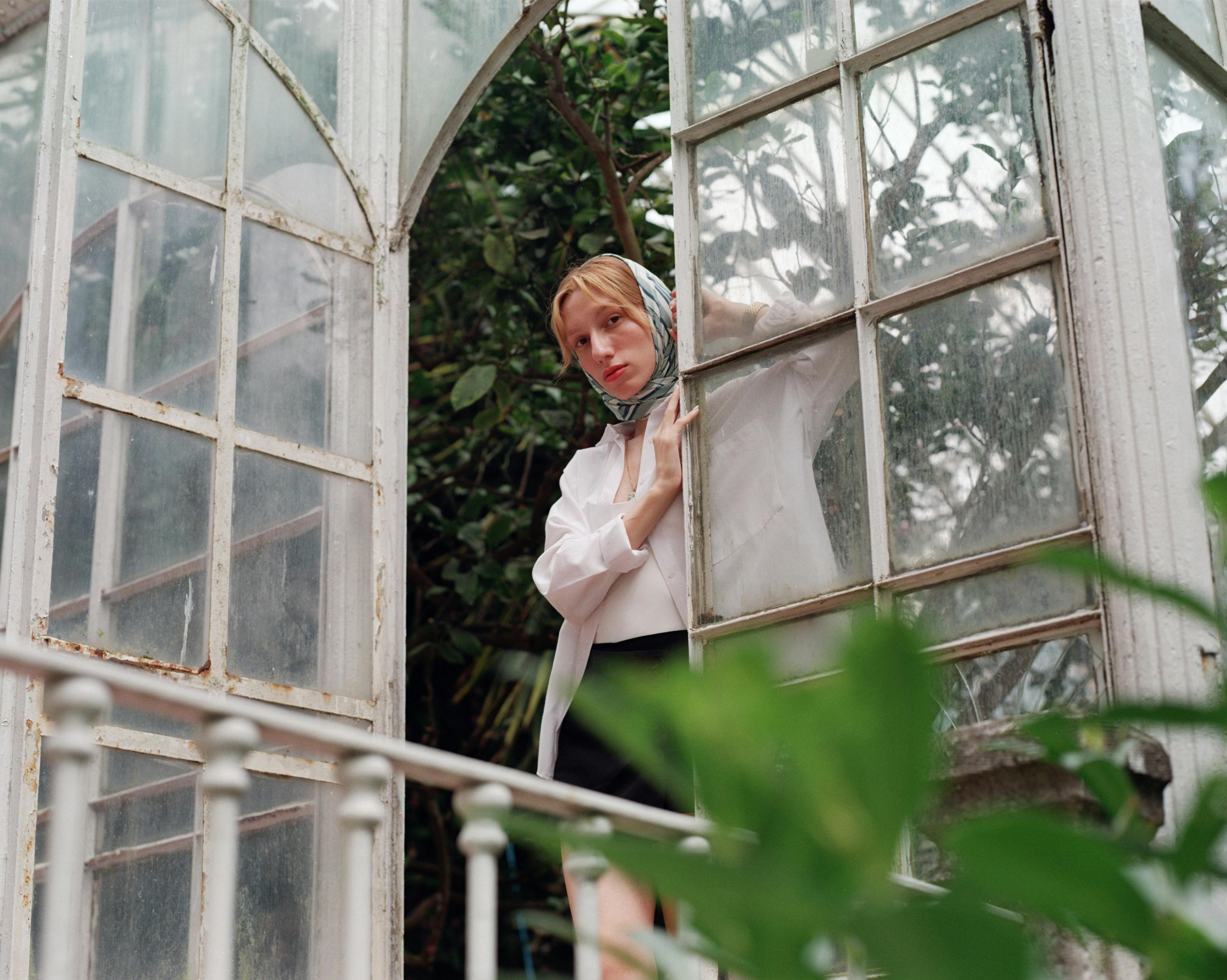 Woman gazing out a greenhouse window amidst lush greenery.