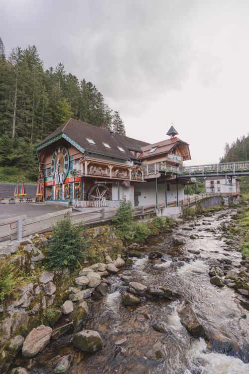 Free Scenic view of traditional German building by a flowing river with lush forest backdrop. Stock Photo