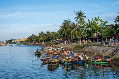 Free Colorful boats along palm-lined riverside in Hoi An, a vibrant scene in Vietnam. Stock Photo