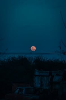 A striking blood moon rises over a dark rural landscape, creating a mysterious evening scene.
