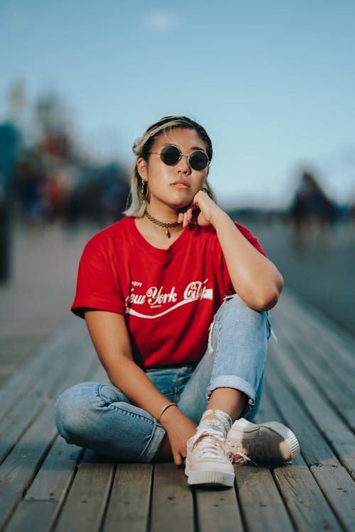 Woman Wearing Red Crew-neck T-shirt and Blue Jeans Sitting on Wooden Floor
