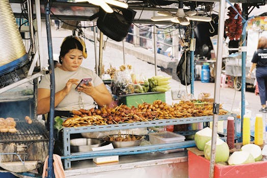 Street vendor grilling skewers at a busy outdoor market, using a smartphone.