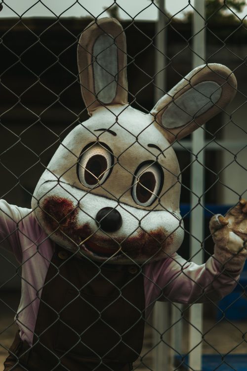 Free Sinister rabbit costume character standing ominously behind a chainlink fence, evoking a spooky atmosphere. Stock Photo