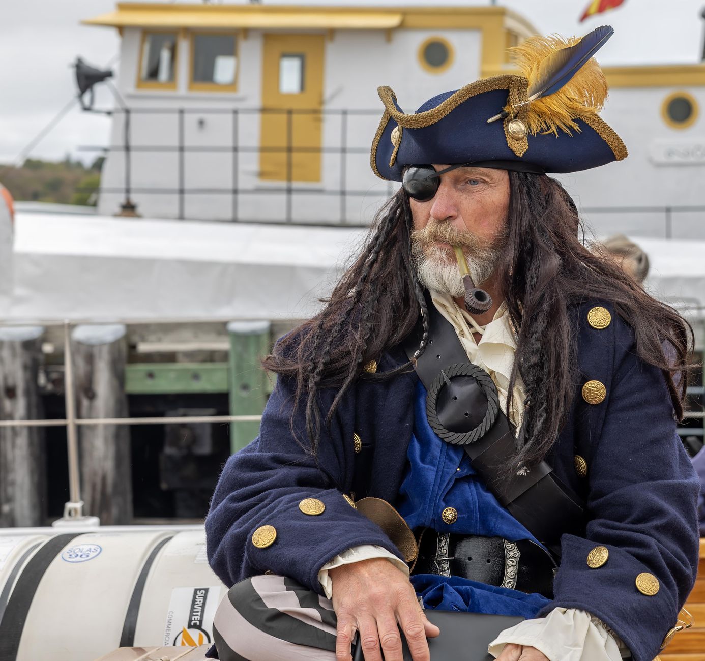 Pirate in costume with eye patch and hat sitting by a boat docked at a harbor.