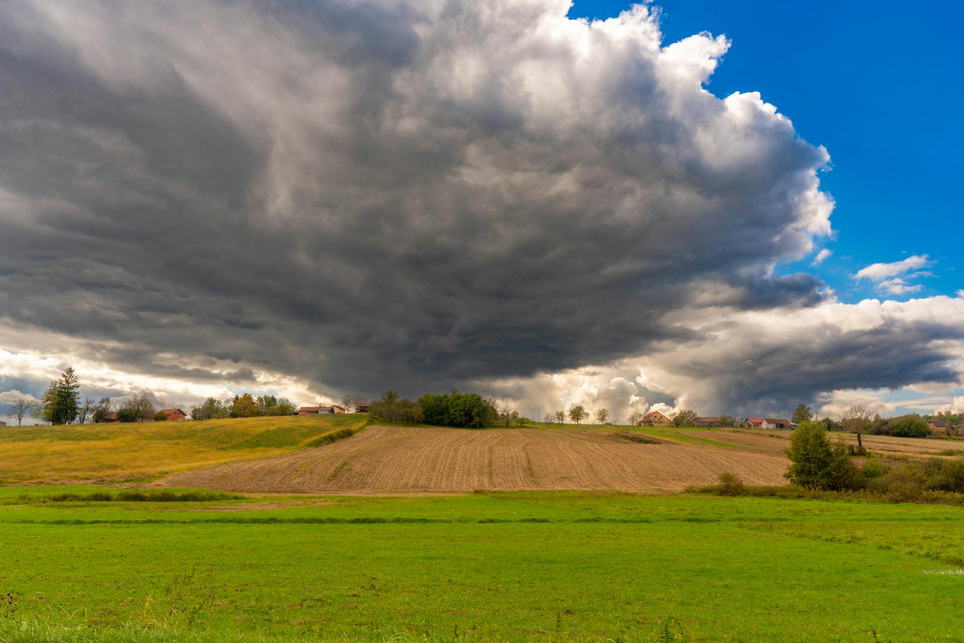 A picturesque rural landscape with dramatic storm clouds gathering over green fields and farmland.