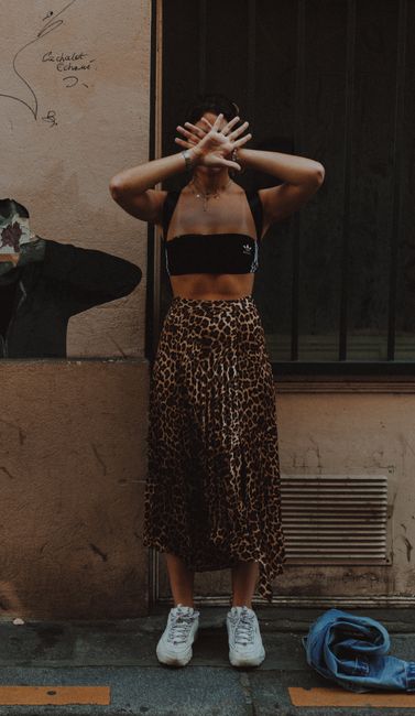 Stylish woman poses with arms raised in Paris alley. Leopard print skirt and crop top enhance urban aesthetic.