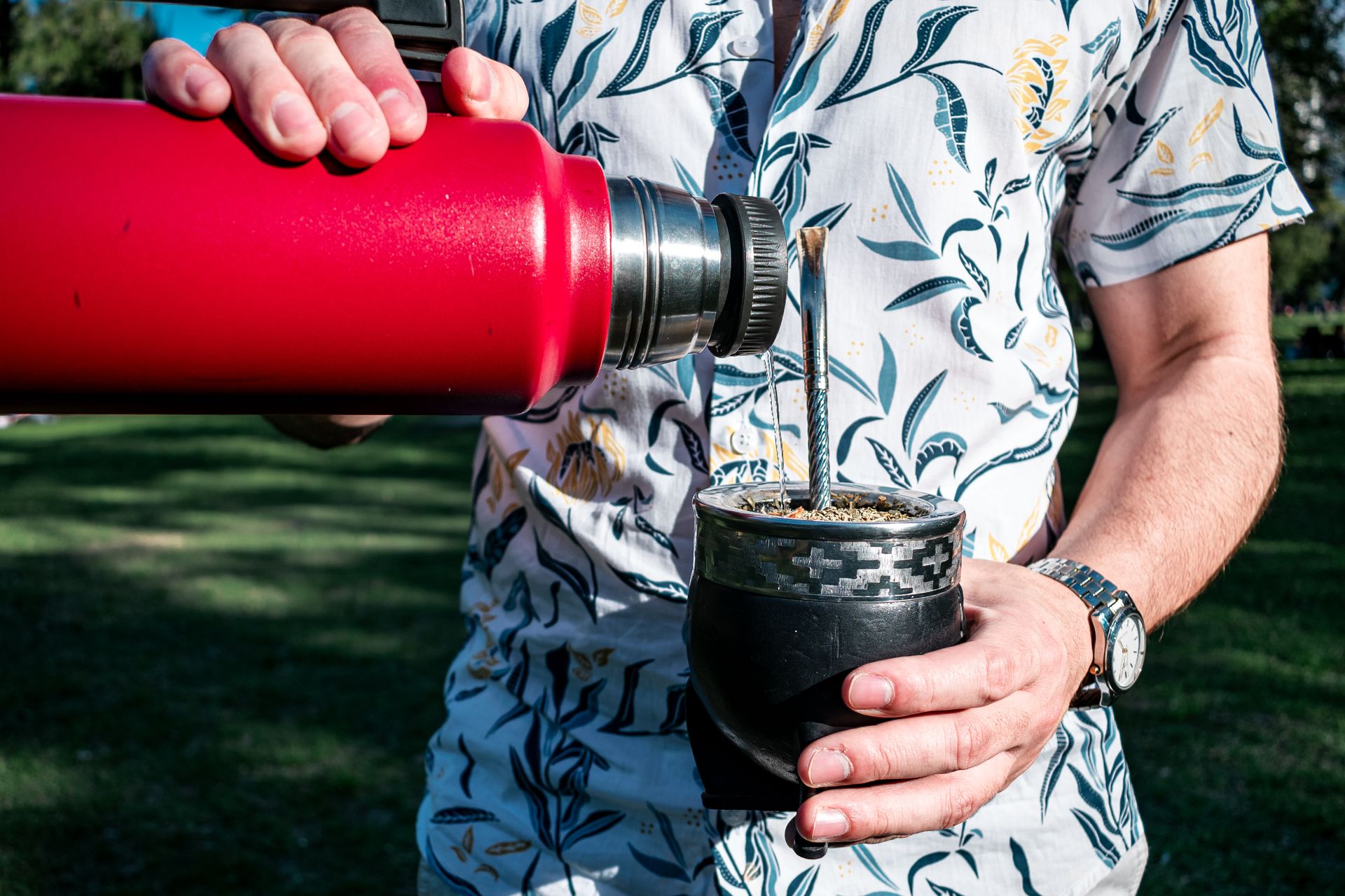 Man pouring hot water from a thermos into a mate cup in a sunny park in Buenos Aires.