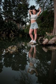 A woman stands on a rock by a reflective pond, surrounded by lush greenery.