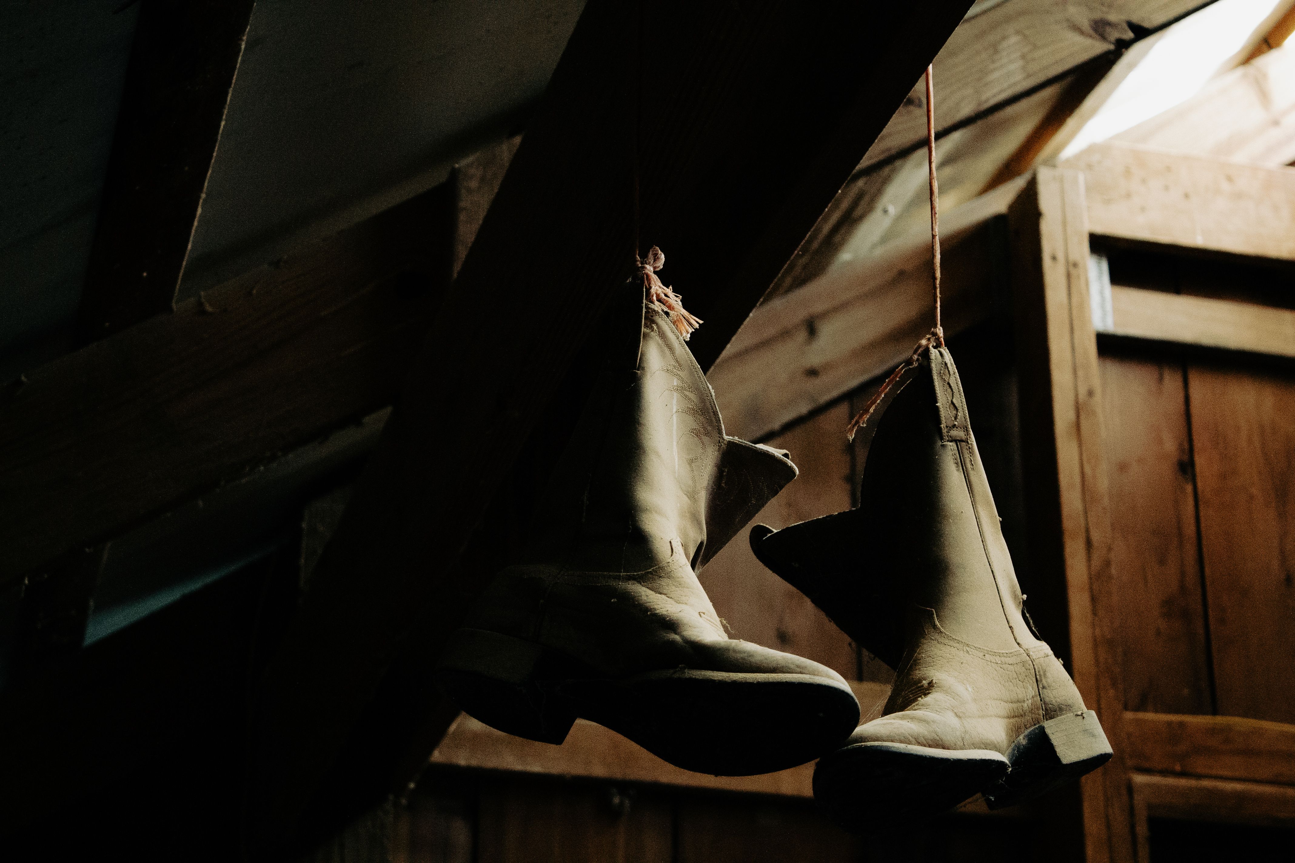 Old cowboy boots hanging in a dimly lit rustic barn setting.