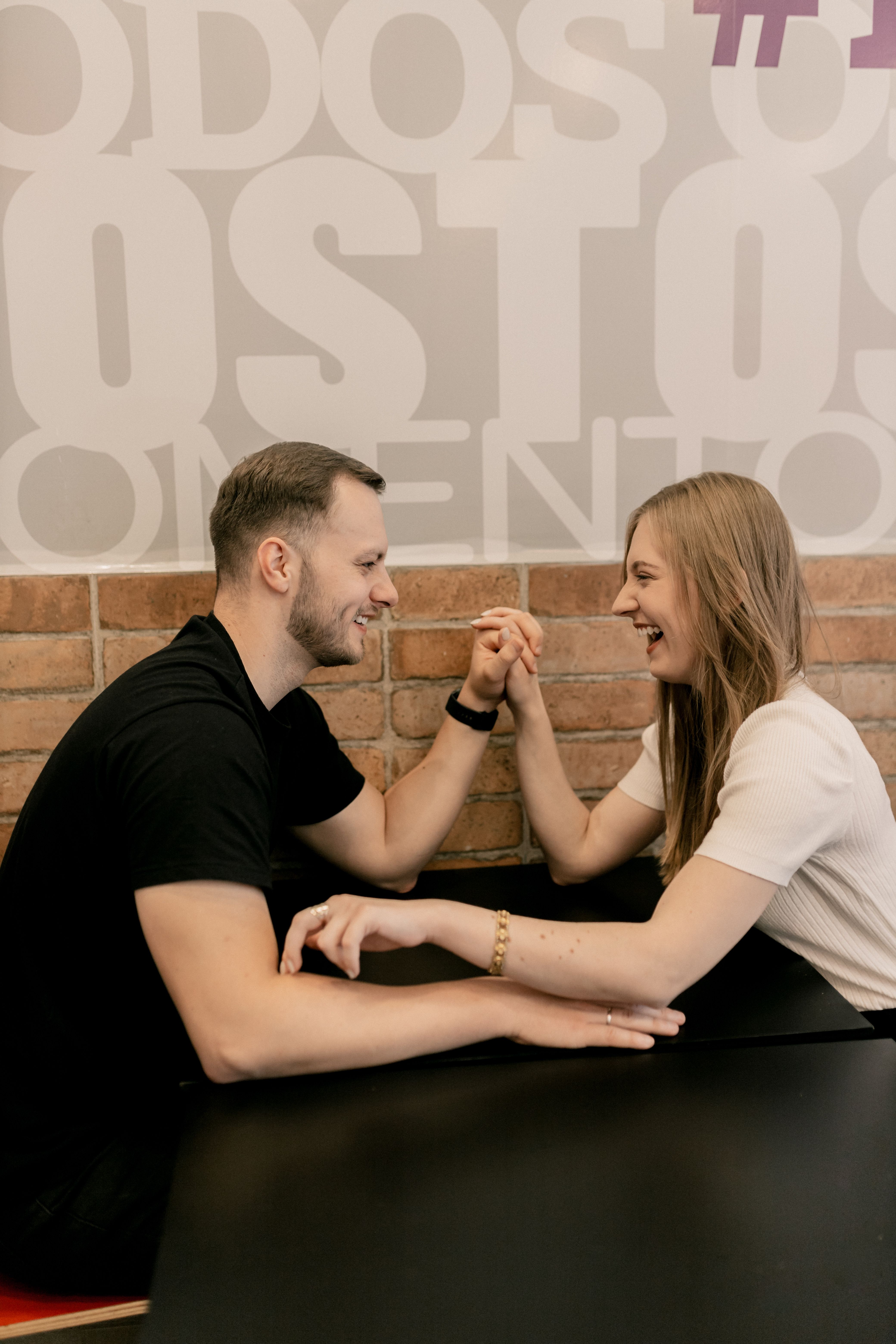 Smiling couple enjoying an arm wrestling match in a cozy café setting.