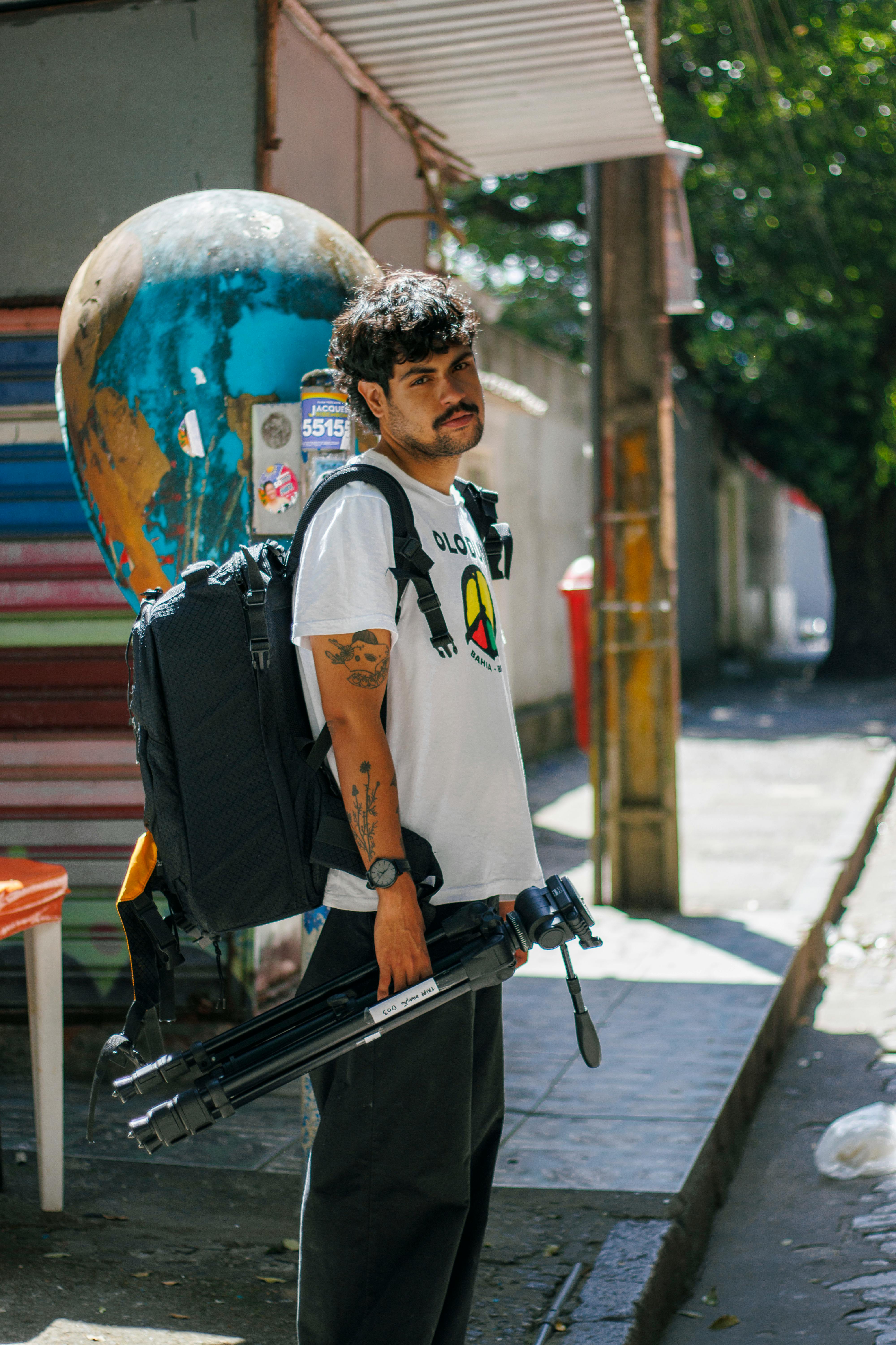 Photographer carrying tripod and backpack in an urban setting on a sunny day.