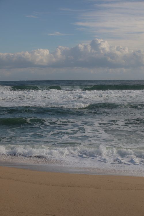 Free Calm ocean waves rolling onto a sunlit sandy beach under a clear sky. Stock Photo