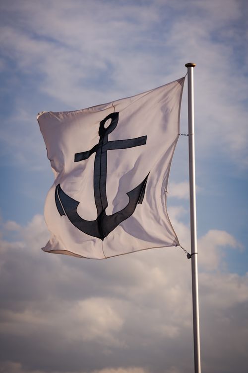 Free A white flag with a black anchor symbol waving against a clear sky, symbolizing nautical themes. Stock Photo