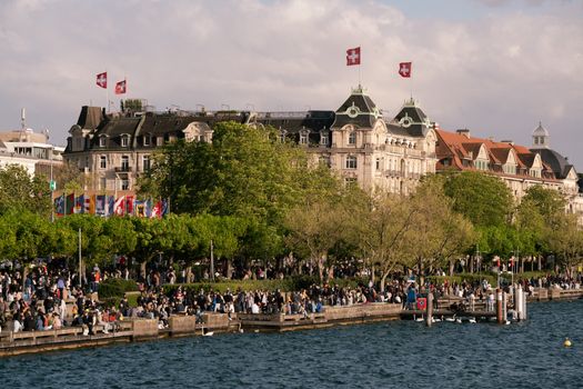 A bustling scene at Zurich's lakefront with crowds and historic buildings under a sunny sky.