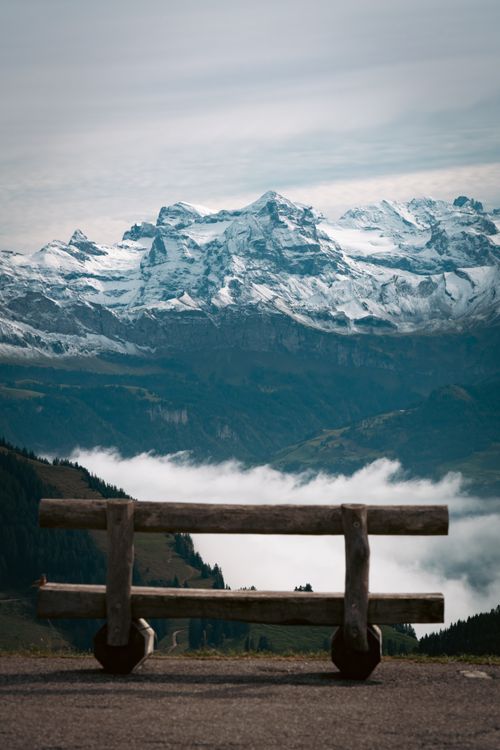 Free Peaceful mountain landscape featuring a rustic wooden bench and snow-capped peaks under a cloudy sky. Stock Photo