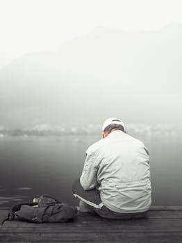 A person in a white jacket sits alone by a misty lakeside, facing the calm water.
