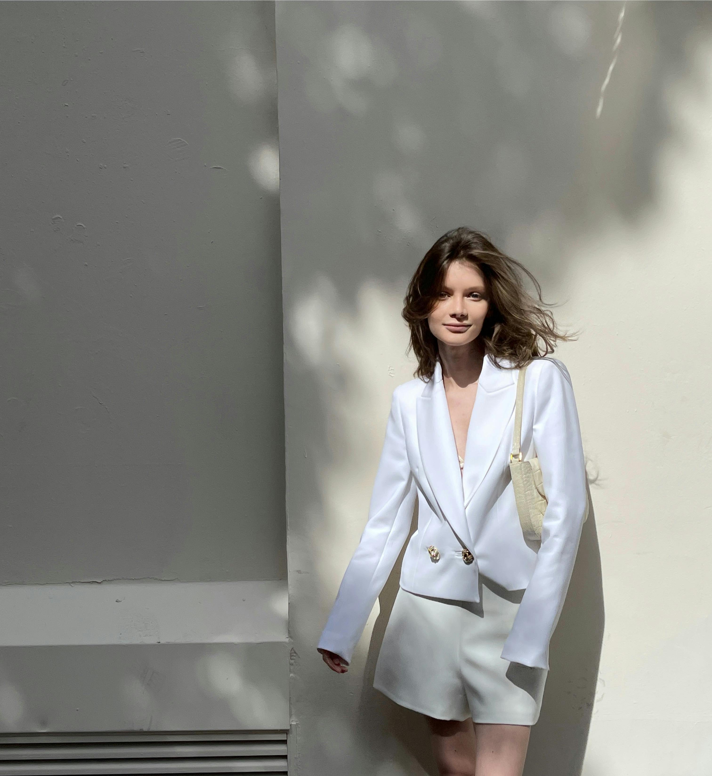 Elegant woman in white blazer and shorts poses against a sunlit wall.