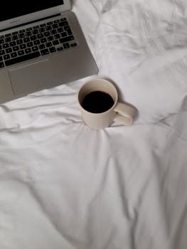 A calming morning scene with a laptop and coffee cup on a white bedspread, perfect for remote work vibes.
