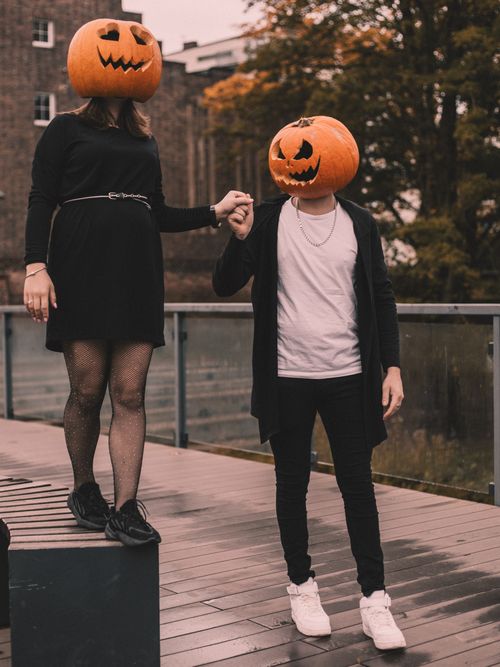 Free A couple dressed in black with pumpkin masks holding hands on a wooden deck, embracing Halloween spirit. Stock Photo