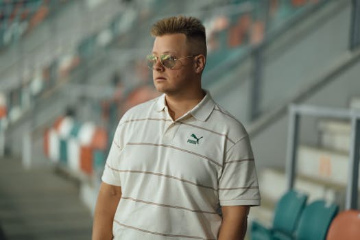 Stylish young man wearing sunglasses and casual attire, standing in a stadium.