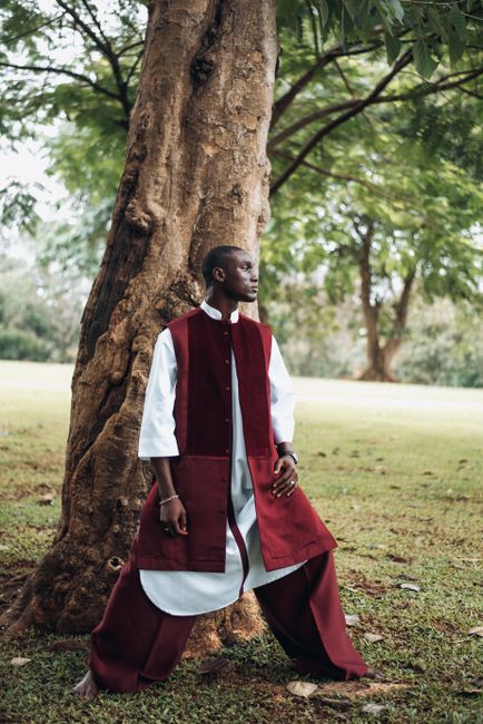 Man in red and white traditional attire leaning on a tree in a lush park setting.