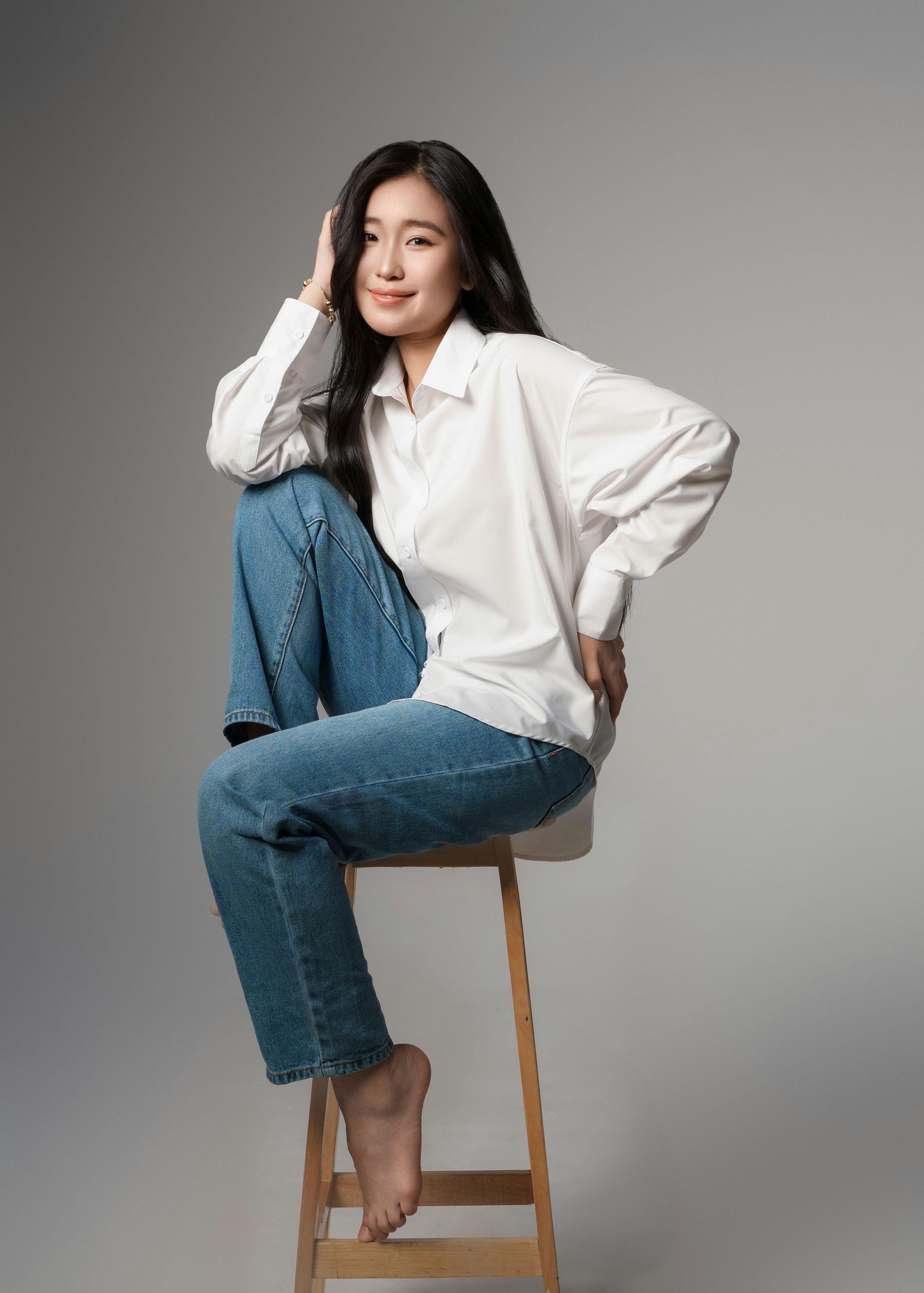 Young woman in casual attire poses confidently seated on a stool in studio.