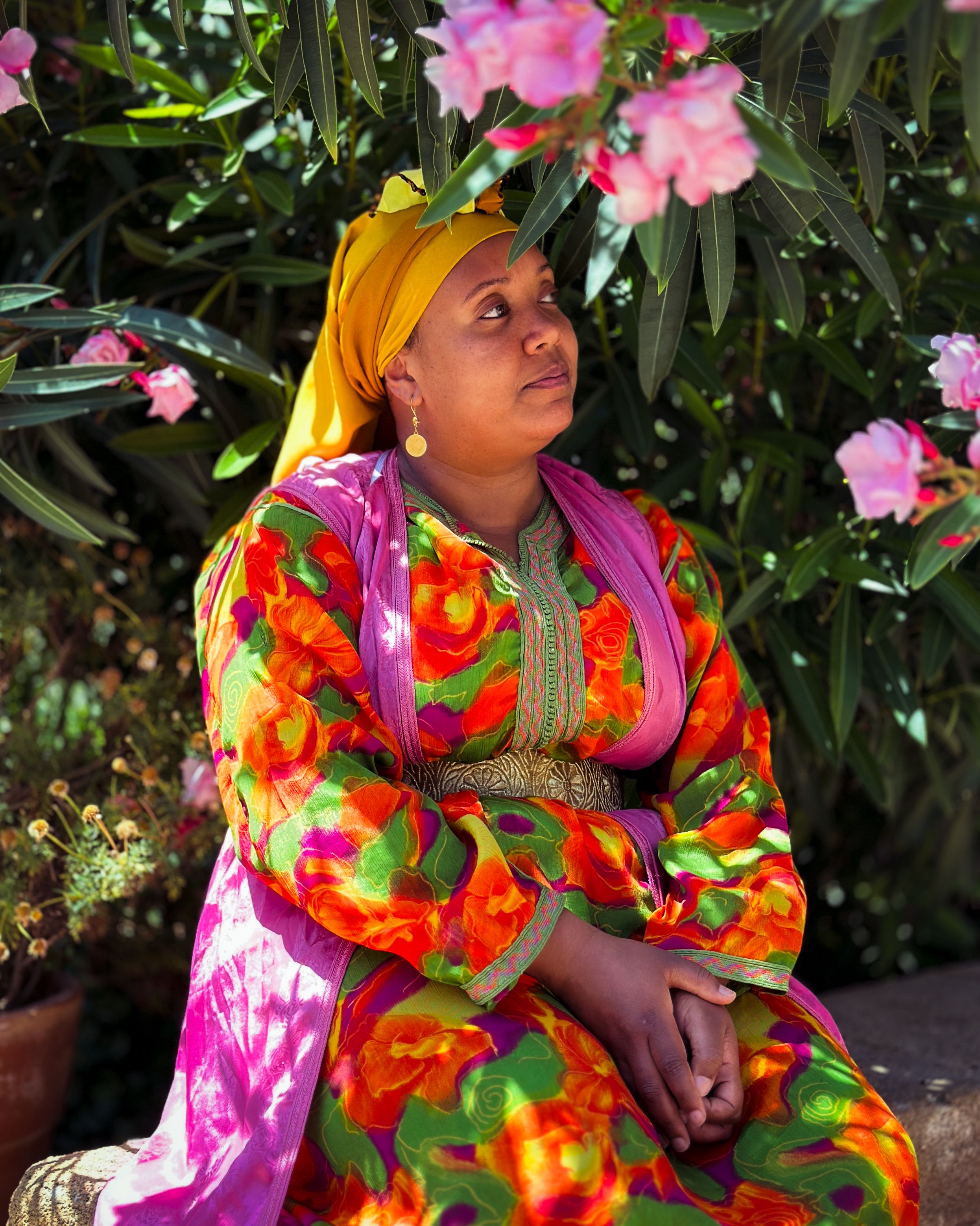 African woman in a colorful floral dress sits outdoors among blooming flowers.