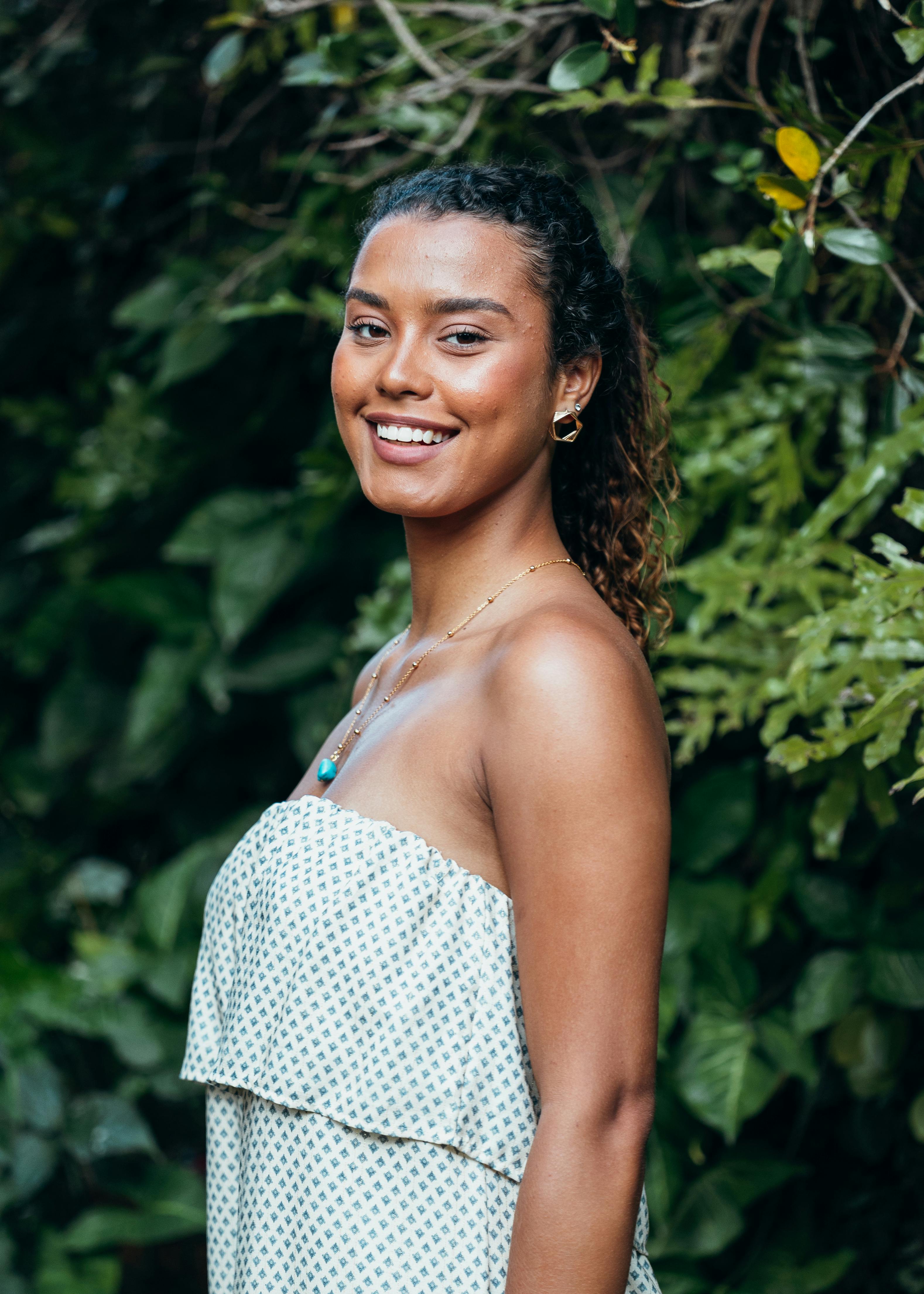 A smiling woman in a strapless dress stands against lush green foliage.