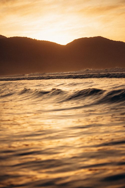 Free A serene ocean scene with gentle waves and mountains silhouetted in a golden sunset. Stock Photo