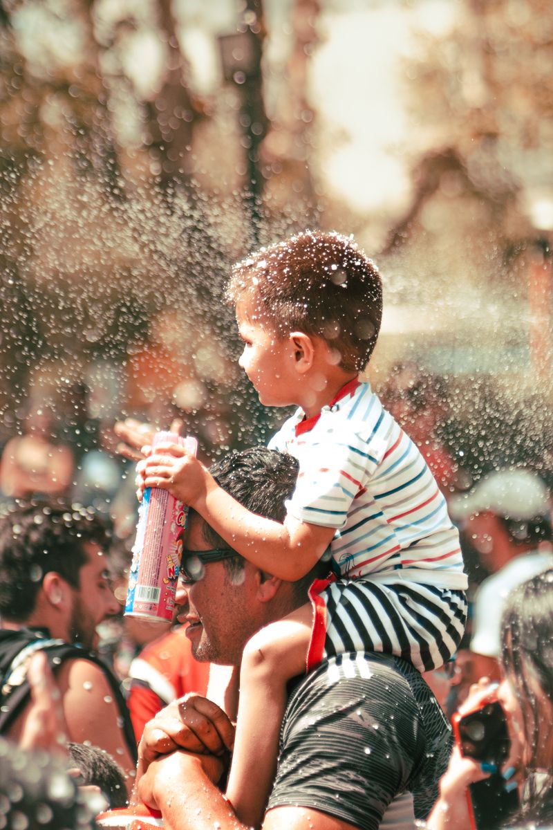 Young child on father's shoulders watching a waterfront event with festive crowd around them