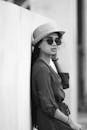 一幅时尚的黑白肖像中，一位戴帽子和太阳镜的女人倚靠在墙上