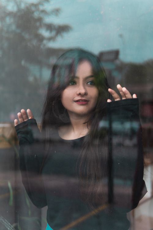 Free Portrait of a woman with long hair posing behind a glass window, capturing a serene and stylish expression. Stock Photo
