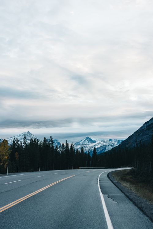 Free A beautiful highway winds through a forest towards stunning snow-covered mountains under a cloudy sky. Stock Photo