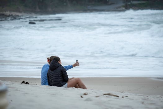 Two friends sitting on a sandy beach taking a selfie near the ocean waves.