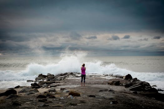 A lone person stands on a rocky jetty as waves crash under a dramatic cloudy sky.