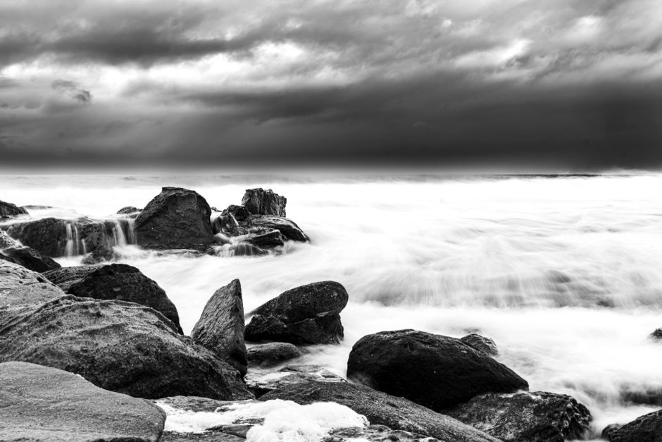 A stunning black and white capture of rugged rocks and crashing ocean waves under stormy clouds.