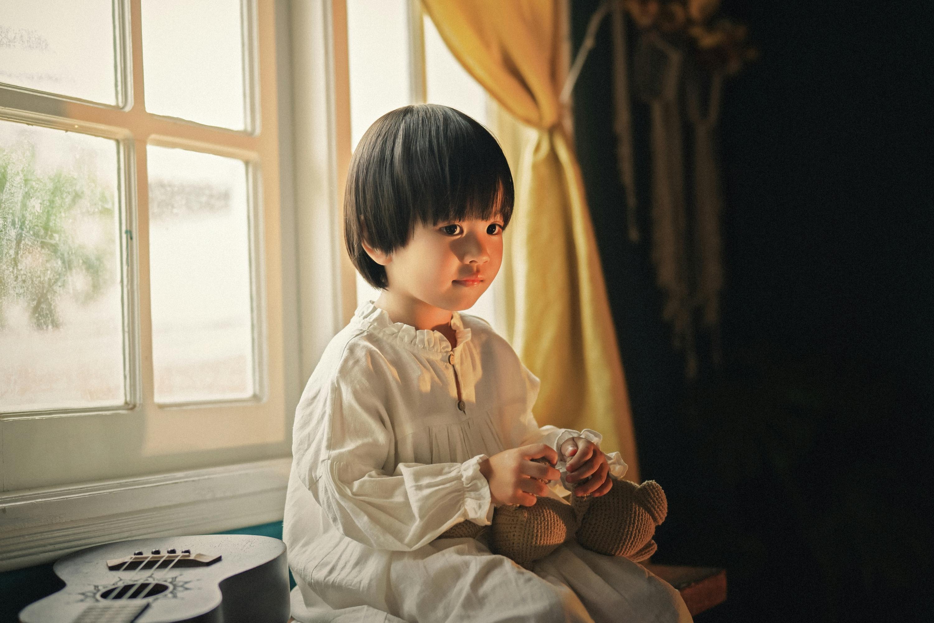 A serene portrait of a child inside, holding a teddy bear by a window.