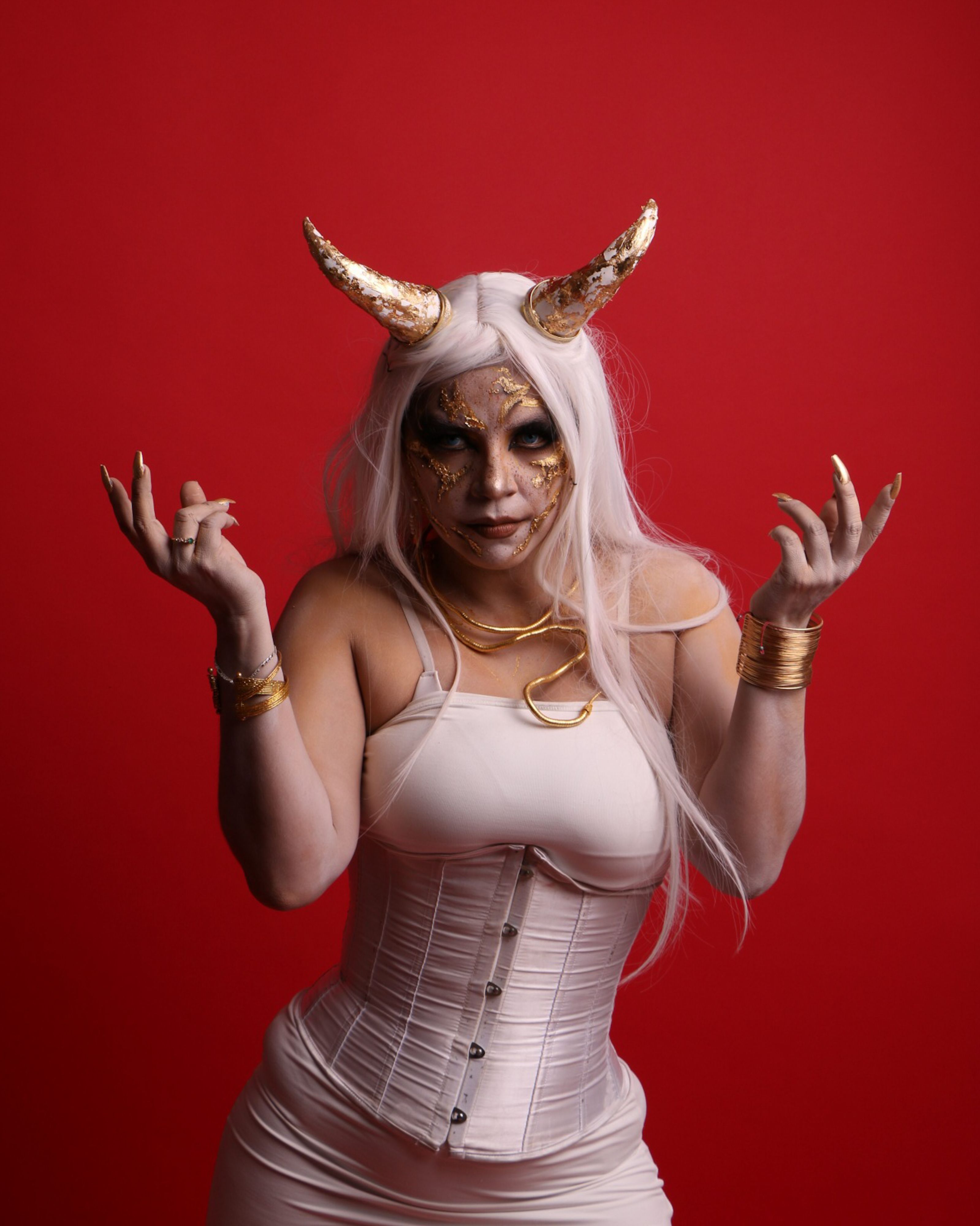 Golden horned figure with intricate makeup and white costume against a red backdrop.