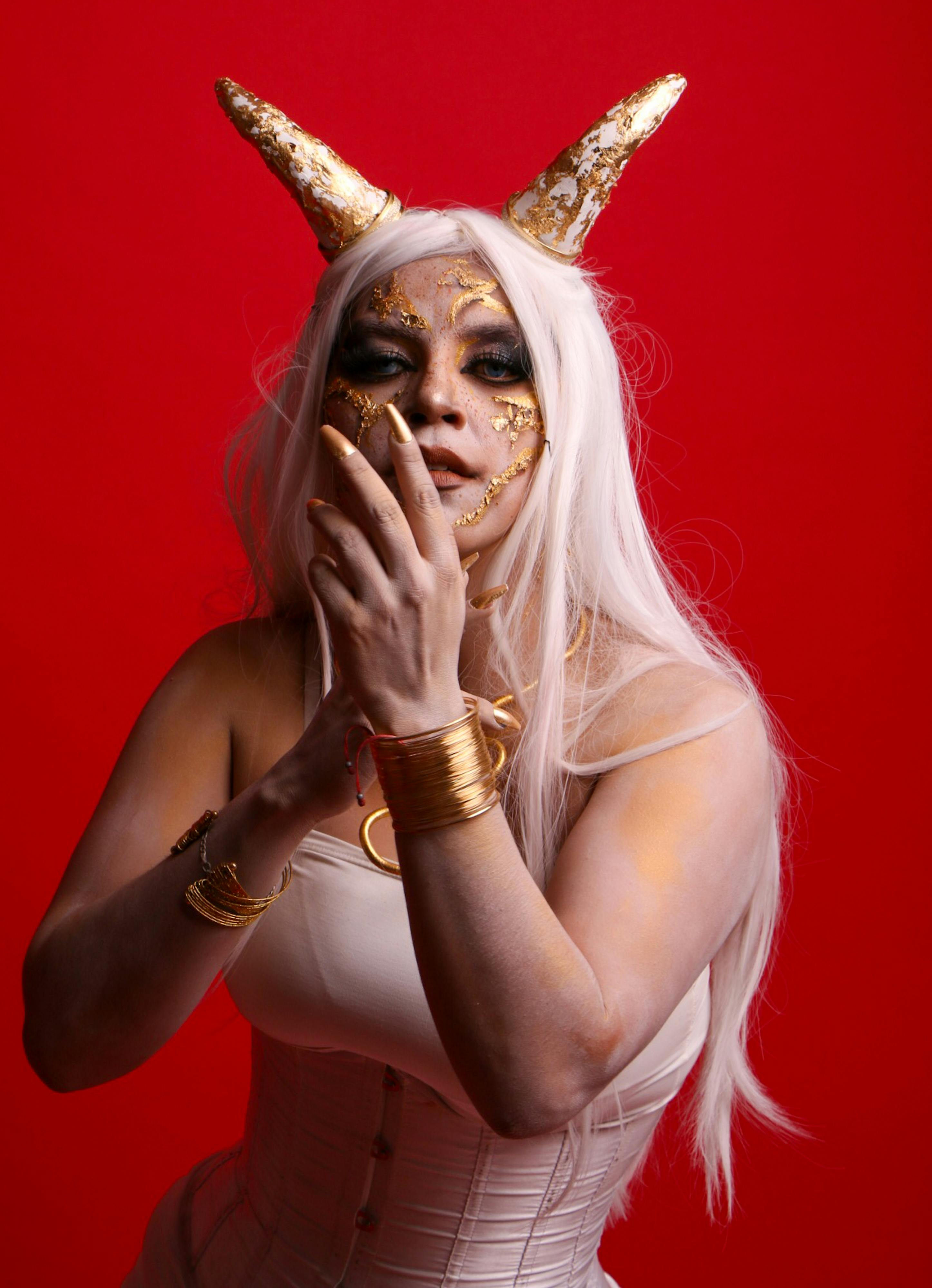 Artistic portrait of a woman in costume makeup with golden horns and a red background.